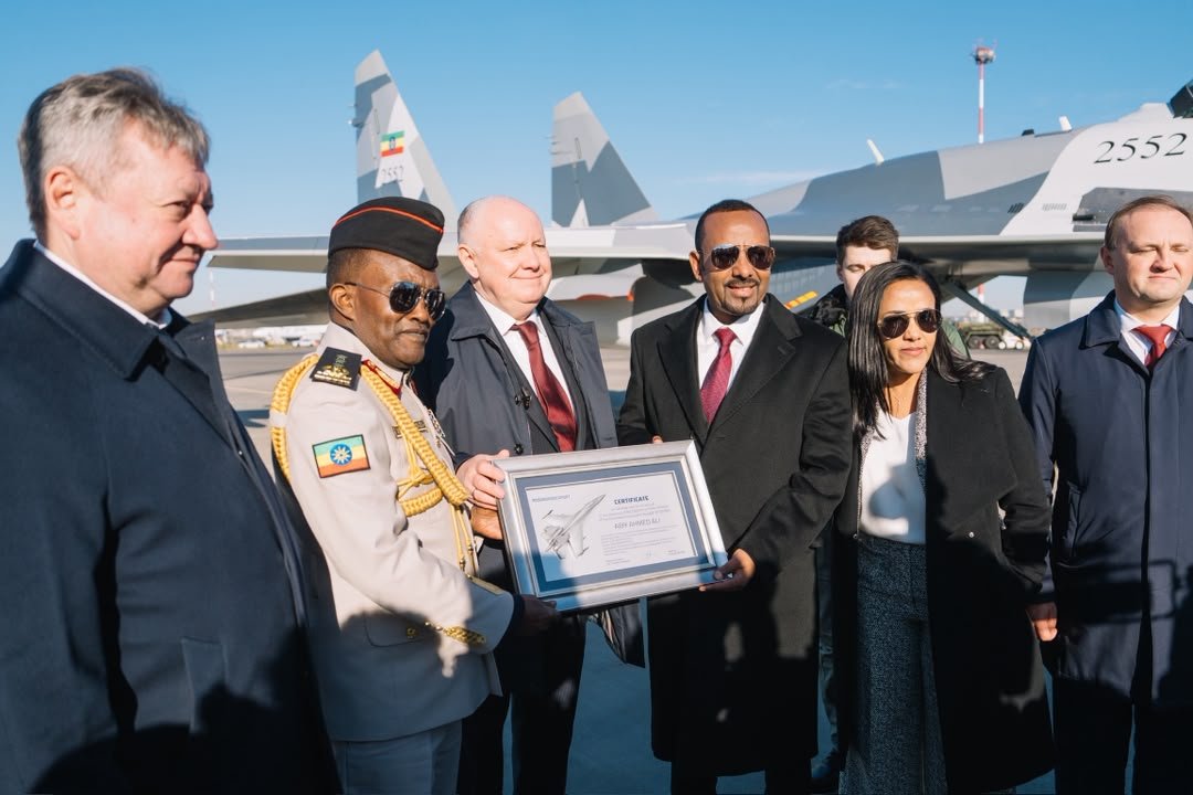 Prime Minister Abiy Ahmed, in sunglasses and a dark coat, holds a framed certificate with a Russian official and an Ethiopian officer in dress uniform, in front of a grey Su-35 fighter jet with the Ethiopian flag on its tail and the number 2552 on its nose.