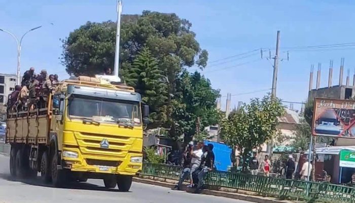 A truck carrying men in military-style uniforms passes bystanders on a town street