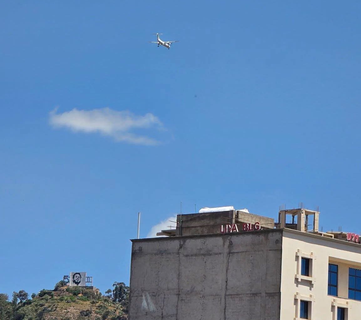 An Ethiopian Airlines propeller plane flies low in a blue sky over a building and a hillside billboard.
