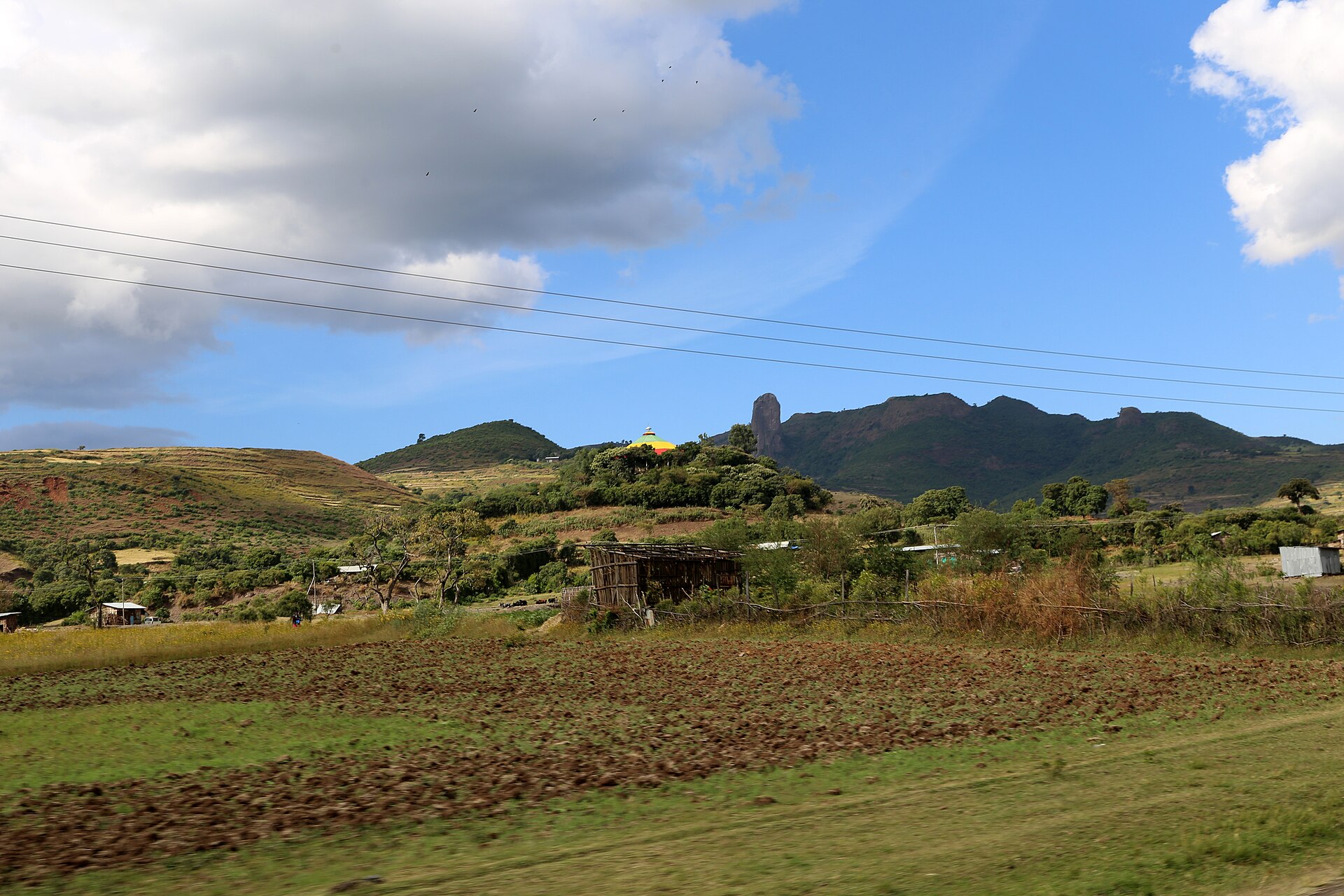 Ploughed fields and scattered trees in front of green hills and a tall rocky outcrop near Addis Zemen, with a yellow-domed church on a hilltop and power lines across a blue sky.