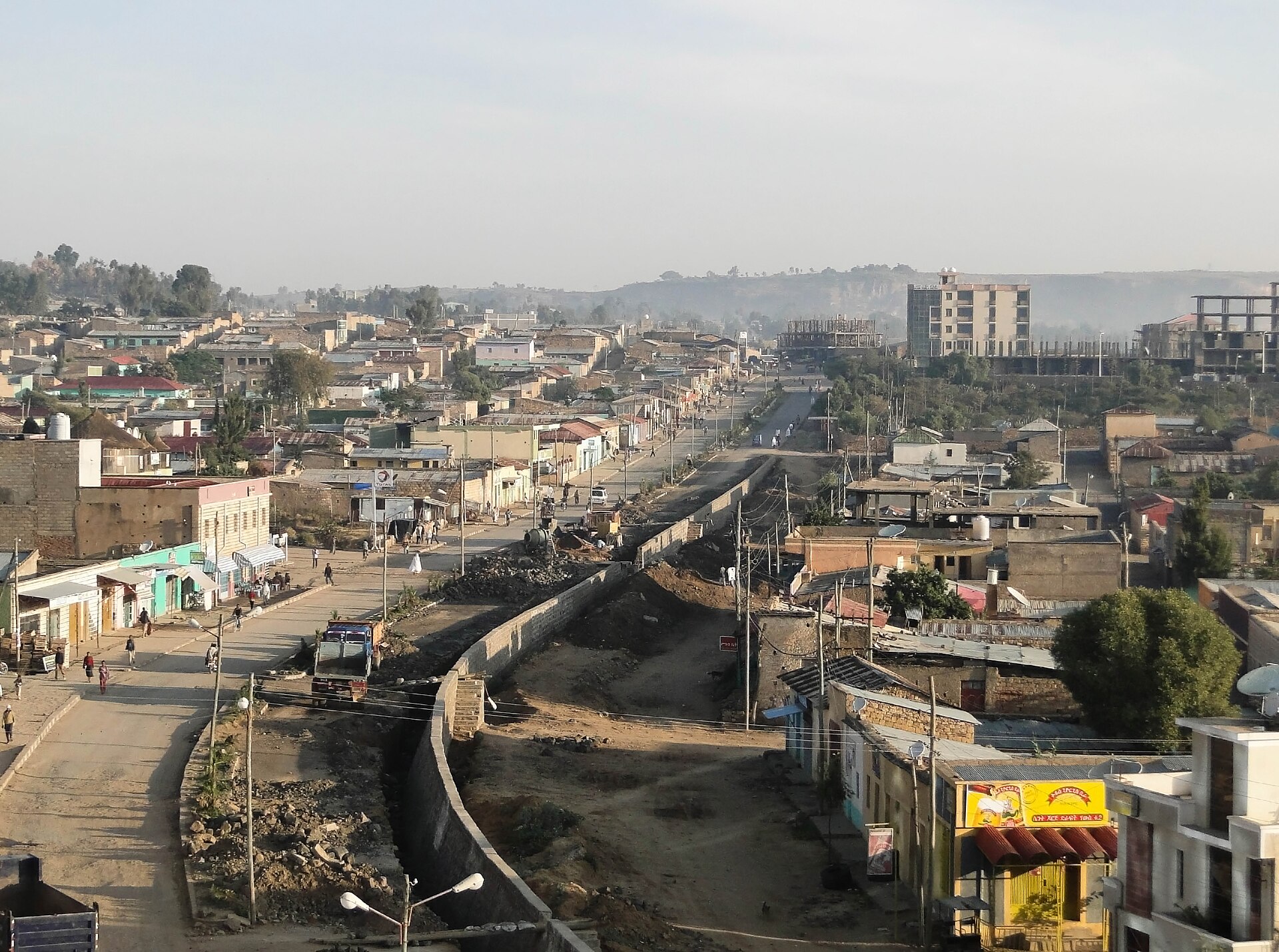 A view down the main road of Adigrat, with low shops and houses on both sides, a half-built multistory building on the right and hazy hills beyond.