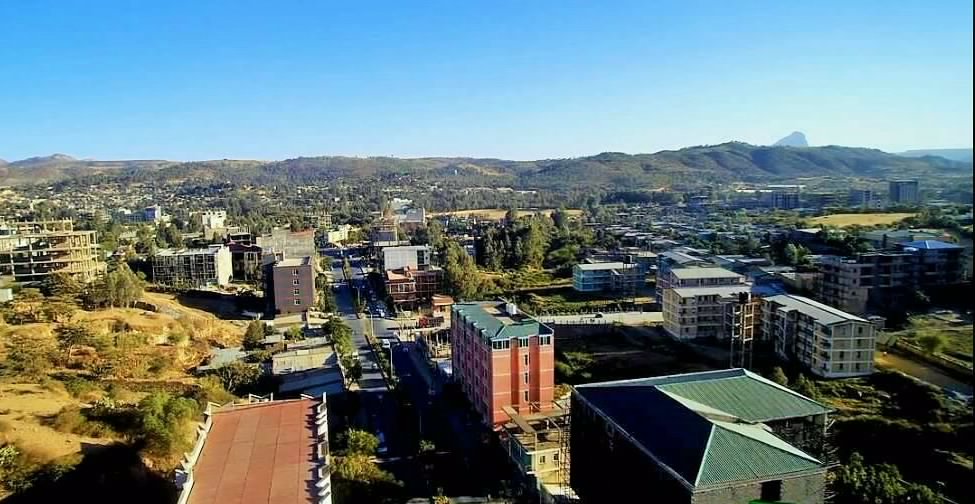 An aerial view of Adwa, with a main road running between mid-rise buildings, trees and hills under a clear sky.
