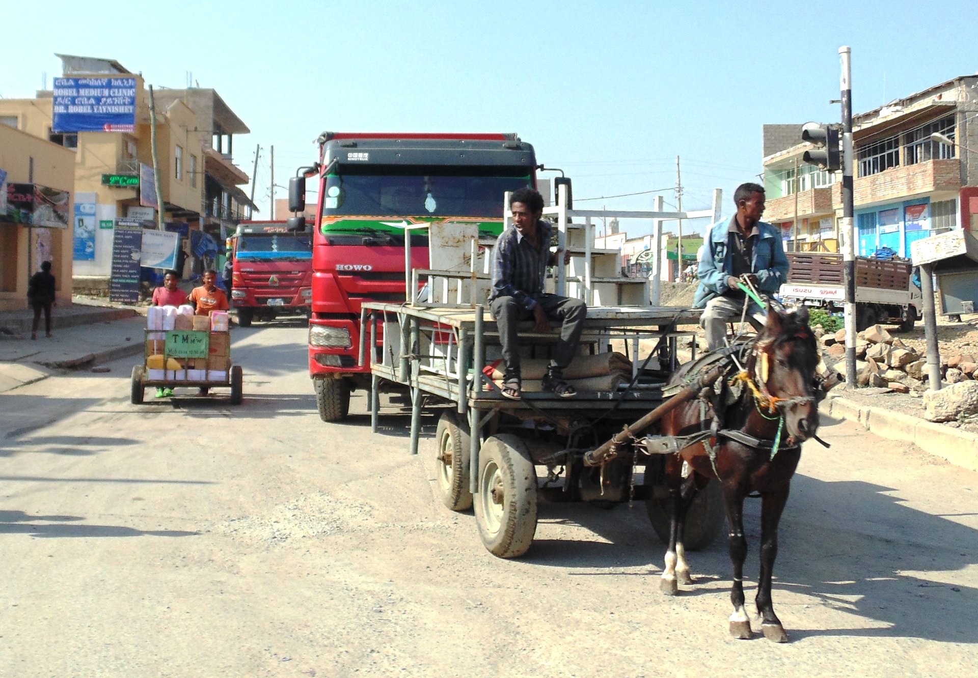 A street in Mekelle with a man driving a horse-drawn cart loaded with furniture, a red truck behind him, and shops and clinic signs along the road.