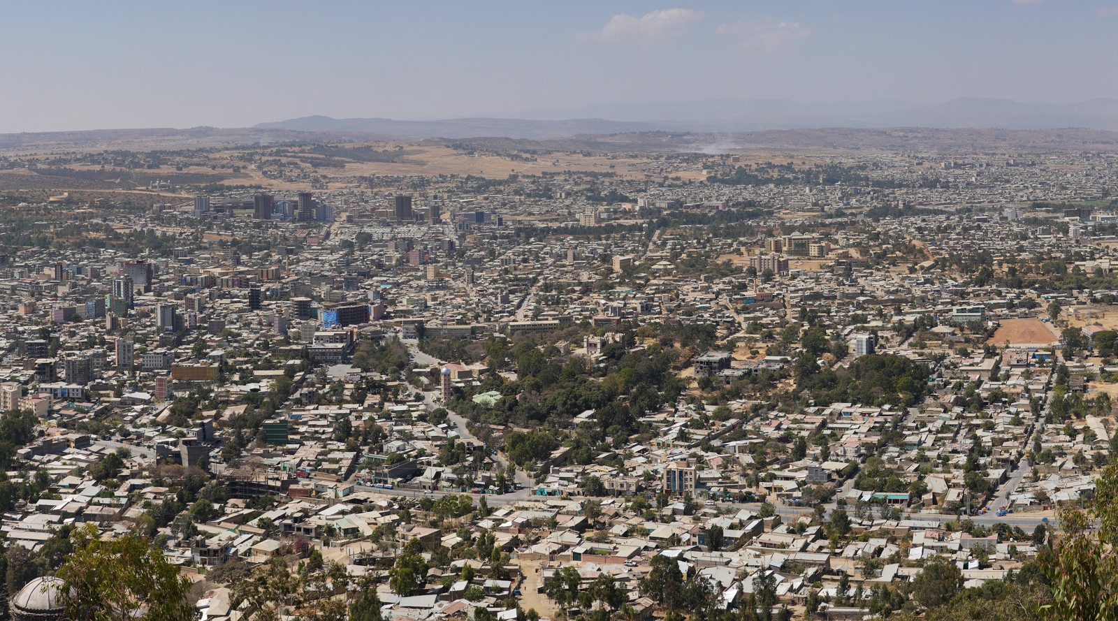A wide view of Mekelle city with low buildings, trees and distant hills under a pale sky.