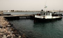 A small white tugboat tied up at a pier in Obock harbor, with the town visible across the water.