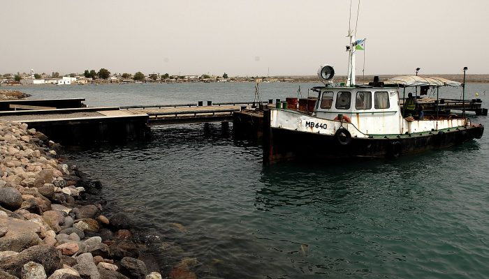 A small white tugboat tied up at a pier in Obock harbor, with the town visible across the water.