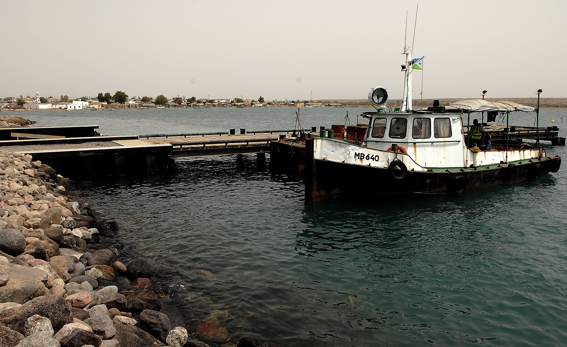 A small white tugboat tied up at a pier in Obock harbor, with the town visible across the water.