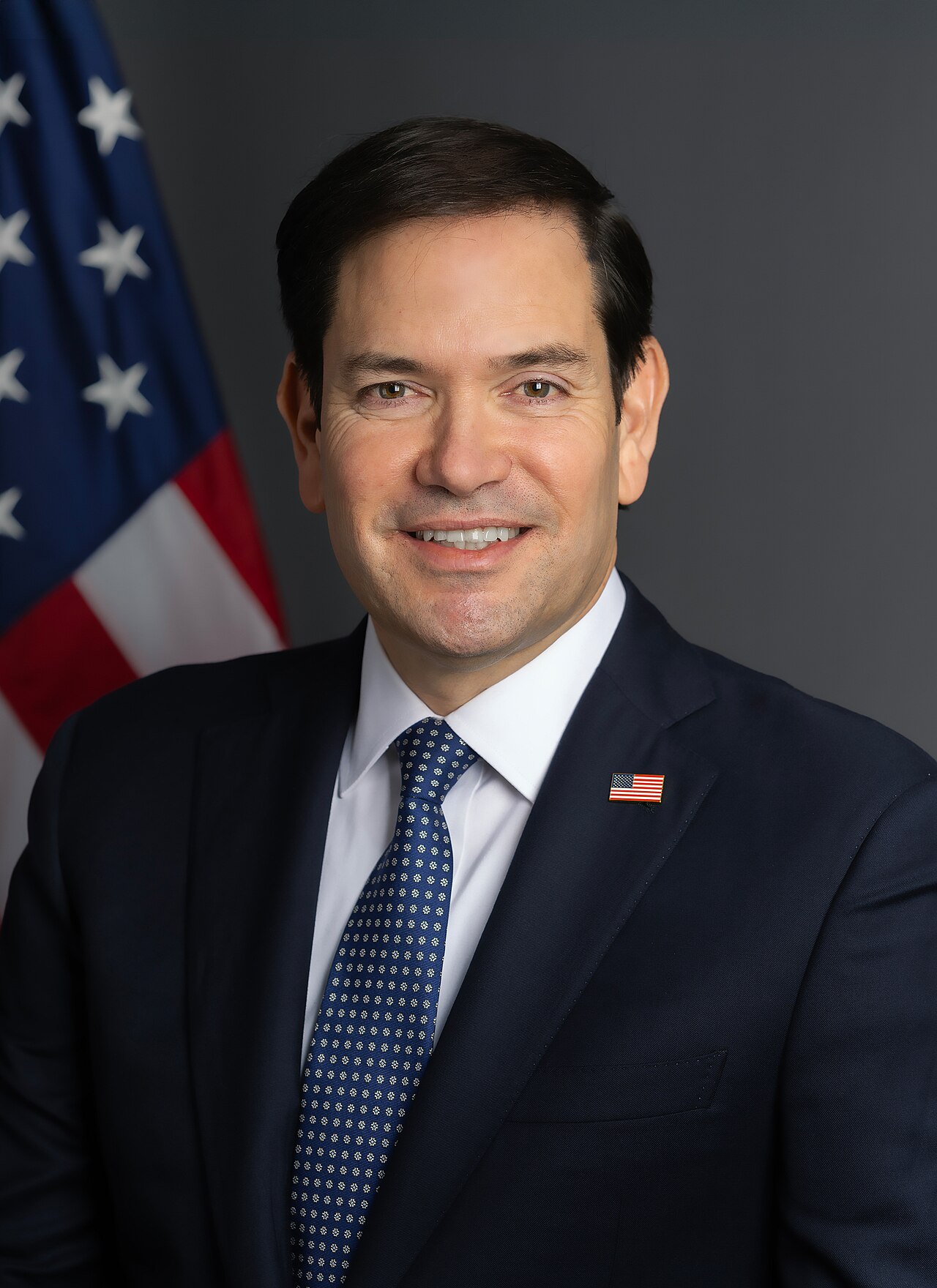 Official portrait of Marco Rubio, smiling, in a dark suit, white shirt and blue patterned tie with an American flag pin, in front of a U.S. flag and a gray backdrop.