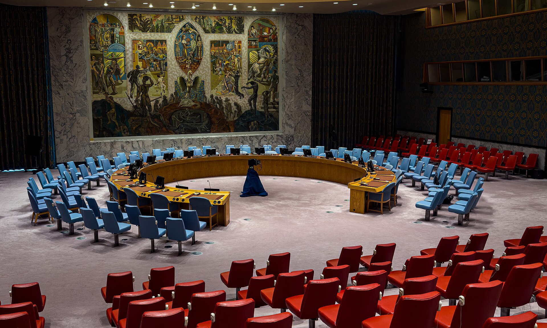 The empty UN Security Council chamber in New York, with its horseshoe table, blue and red seats and a large mural on the far wall.