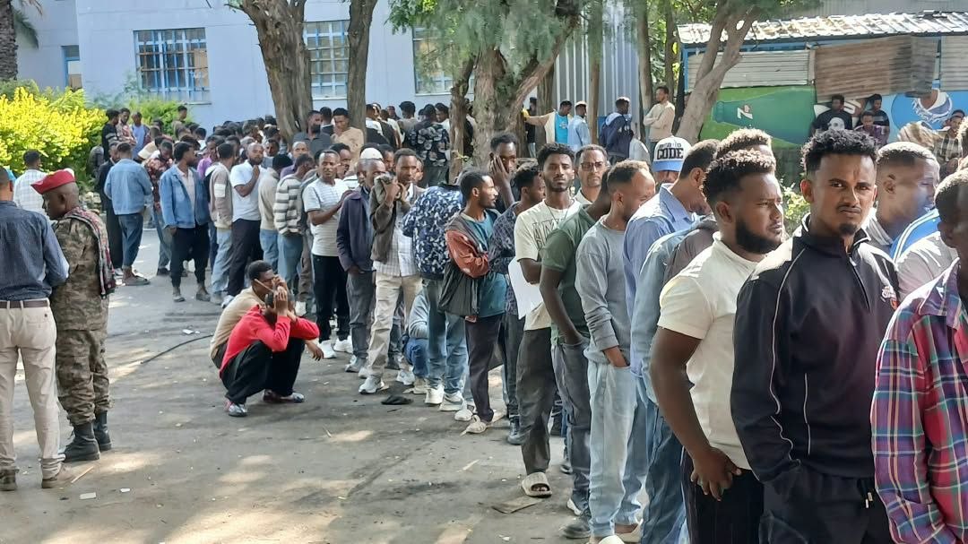 A long line of young men in casual clothes stands along a tree-lined path outside a white building in Mekelle, with a soldier in camouflage and a red beret at the left edge of the frame.