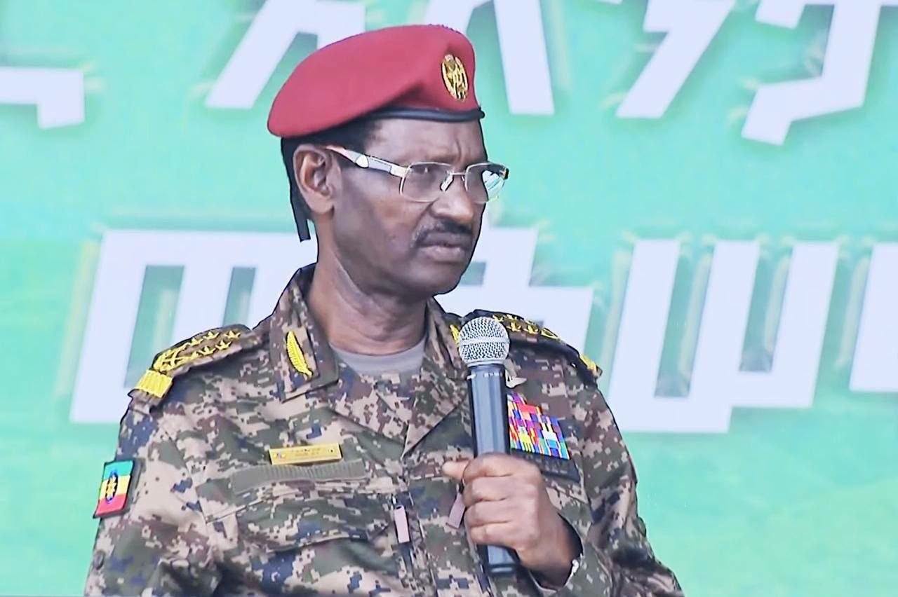 Field Marshal Birhanu Jula, in a red beret, glasses and camouflage uniform with gold shoulder insignia and an Ethiopian flag patch, speaks into a handheld microphone in front of a green backdrop with large white Amharic lettering.