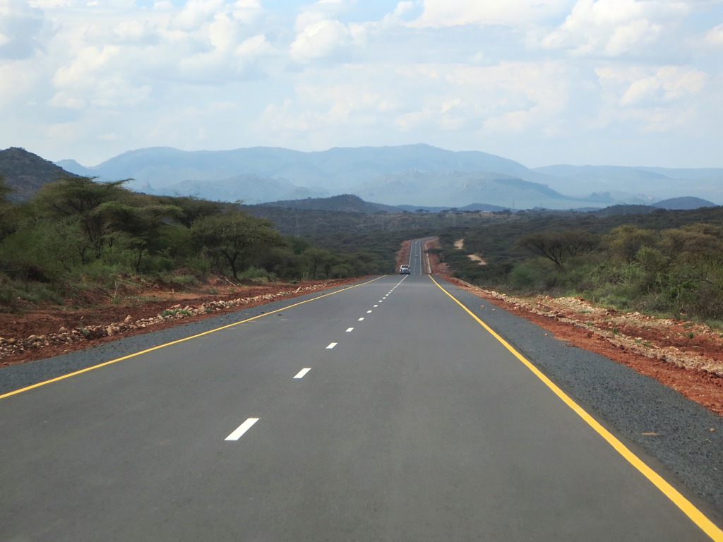 A newly paved two-lane highway with yellow edge lines running straight through green acacia bush toward blue hills, with a single vehicle far down the road, on the route between the Kenyan border at Moyale and Addis Ababa.