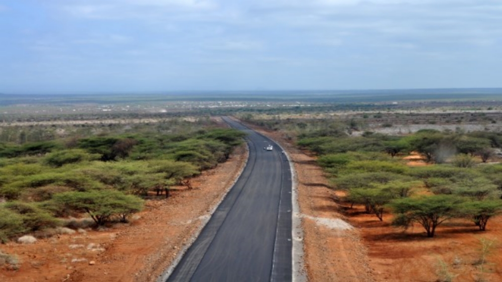 A newly paved two-lane road running straight through flat, dry acacia scrubland toward Moyale on the border between Kenya and Ethiopia, with a single car in the distance under a cloudy sky.