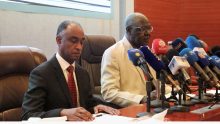 Two Sudanese officials sit at a table behind a row of television station microphones at a news conference in Khartoum.