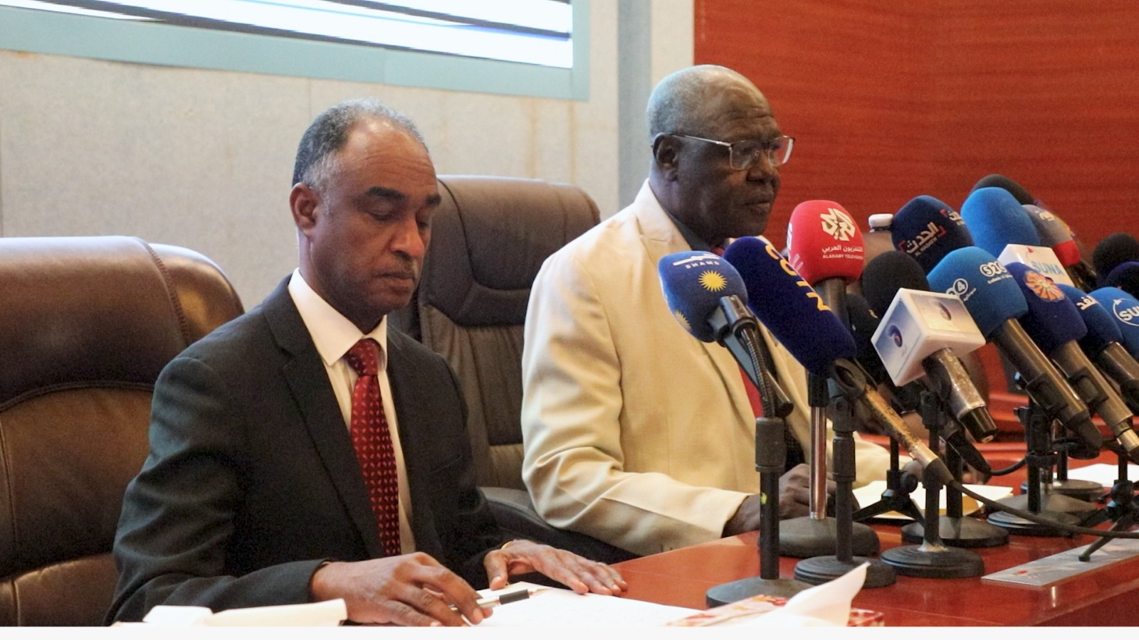 Two Sudanese officials sit at a table behind a row of television station microphones at a news conference in Khartoum.