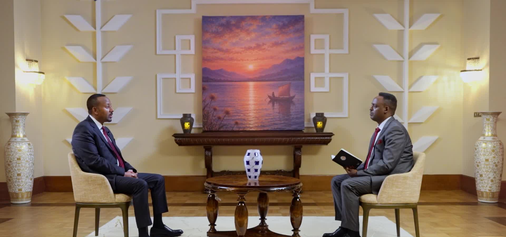 Abiy Ahmed, in a dark suit and red tie, sits in a cream armchair facing an interviewer who holds a notebook, with a painting of a sailing boat on the water hanging on the wall between them.