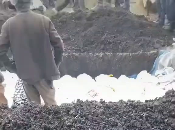 A man in a brown jacket stands at the edge of a long, freshly dug trench in which bodies wrapped in white cloth lie side by side, with piles of dark earth around it.